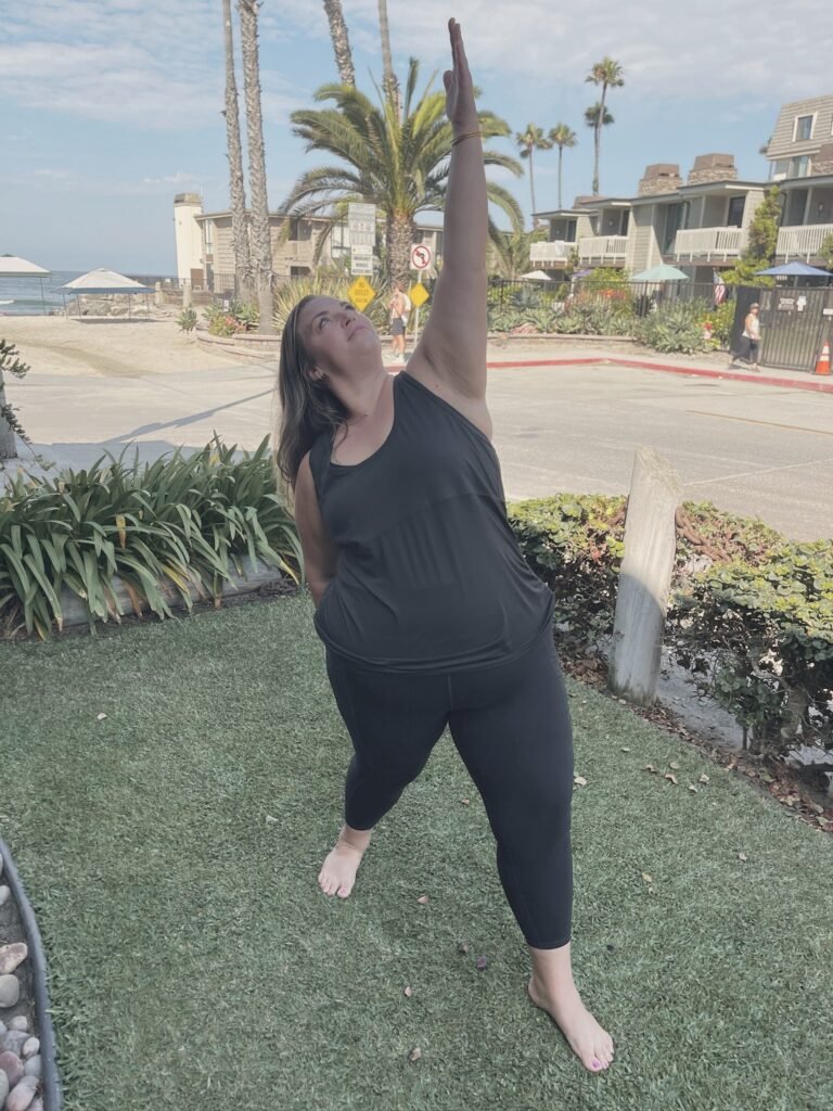 A woman reaching one arm overhead in a side stretch, with the ocean behind her