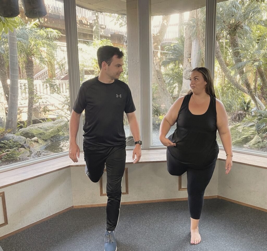 A man and a woman balancing on one leg in front of garden windows