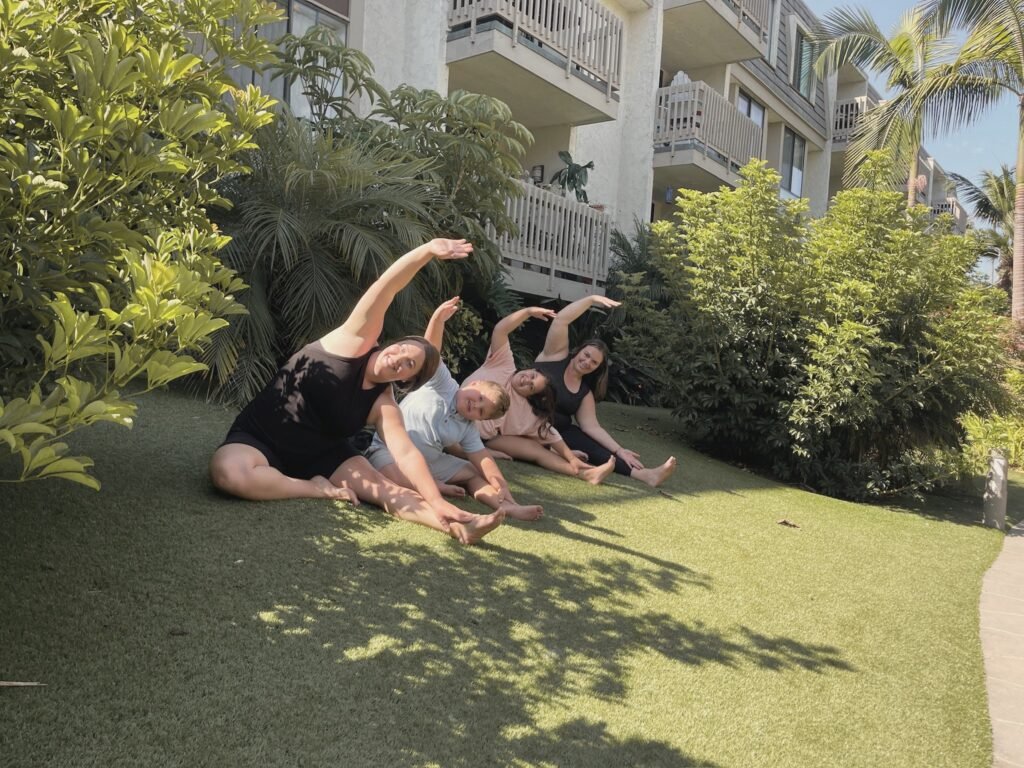 Two women and two children leaning into a seated side bend on the grass